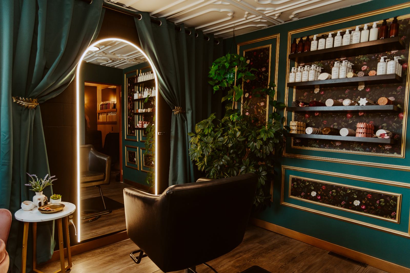 The hair salon space, moss-green walls with gilded paneling, an arched LED mirror, and a leather styling chair surrounded by lush plants.
