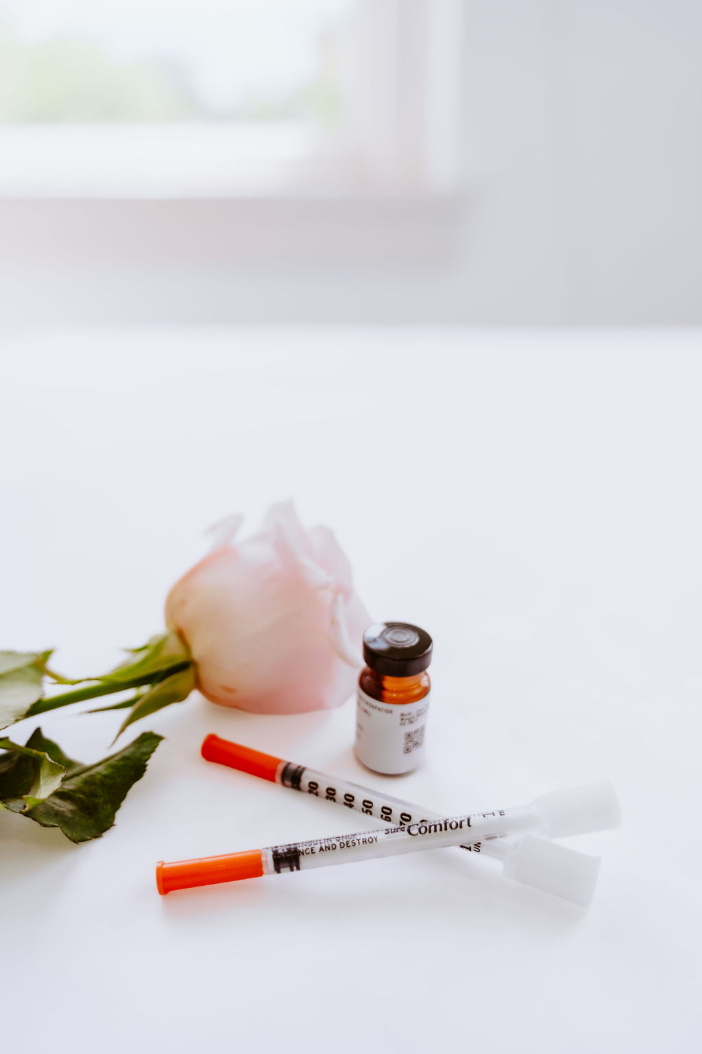 An amber medication vial, two syringes, and a pale pink rose arranged on a white surface