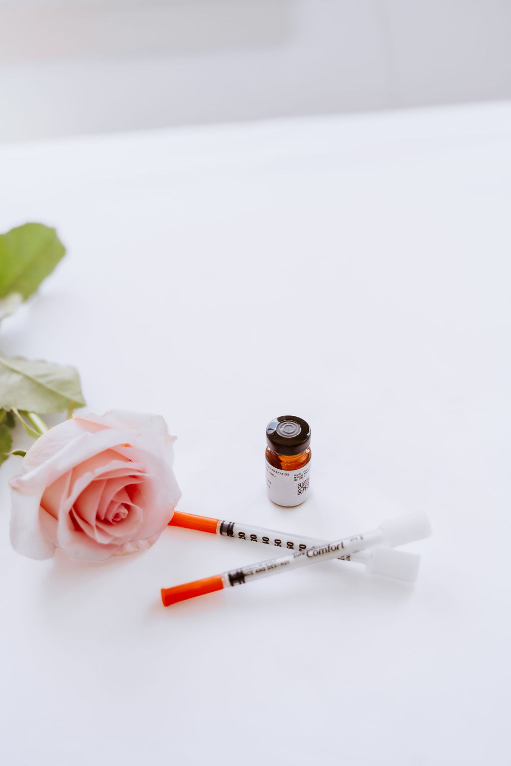 A medication vial and two syringes resting beside a single blush rose on a white surface